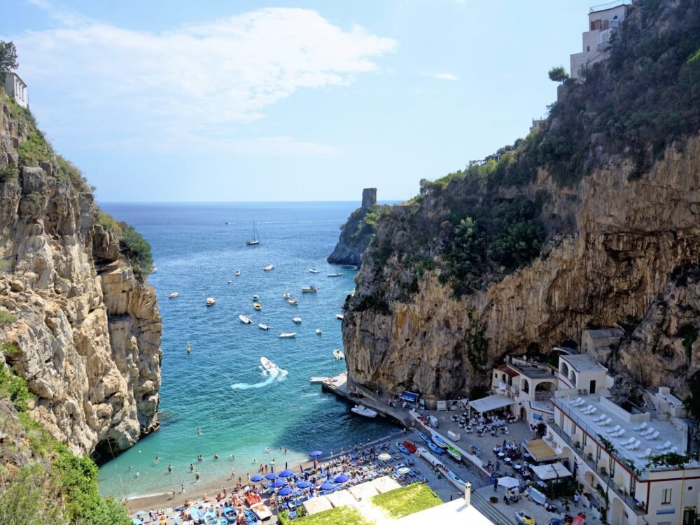 The beach of Marina di Praia, a small beach tucked away between two cliff sides with boats, people in water, sun loungers and beautiful blue water