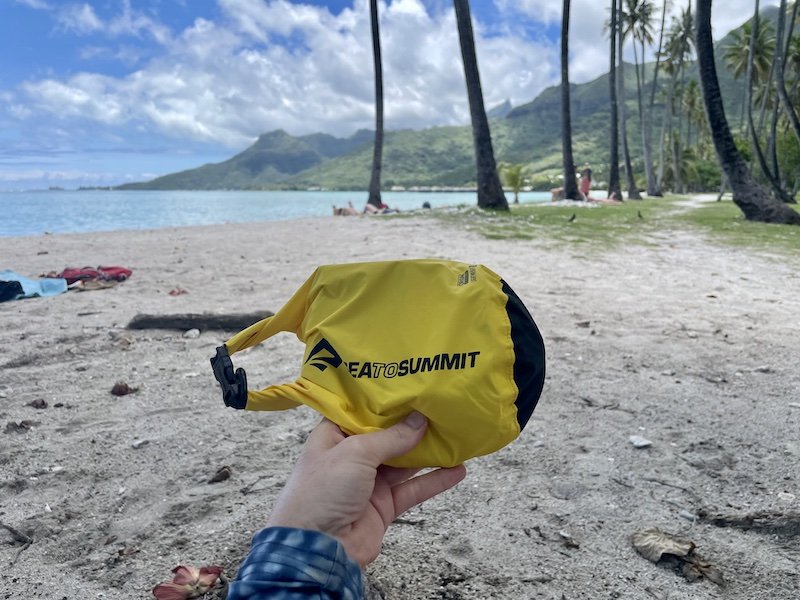 Yellow Sea to Summit dry bag all folded up, in a hand, on the beach with palm trees and blue water and green mountains in background