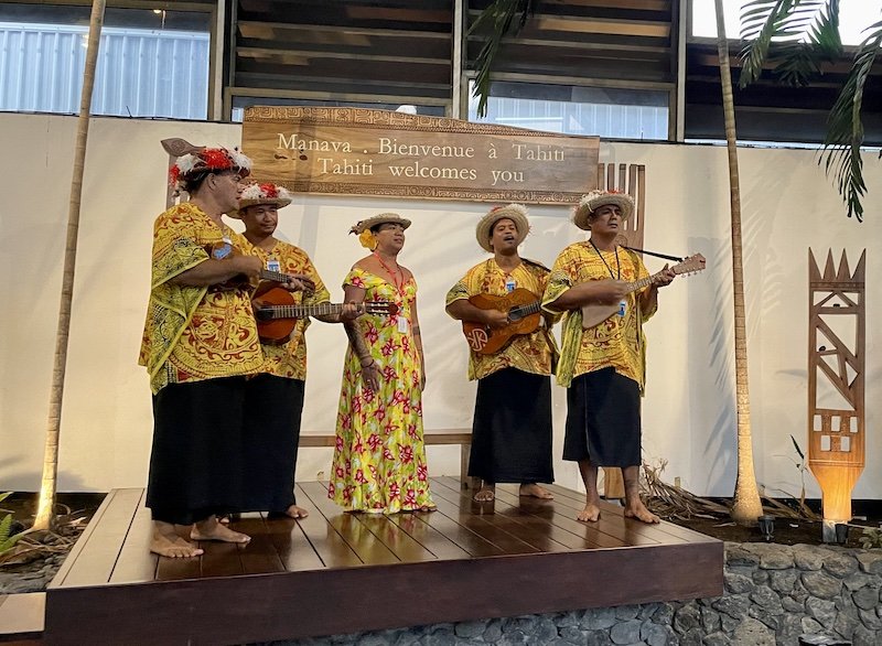 A five-person band of local Polynesians wearing yellow outfits, playing instruments and welcoming arriving guests off the plane as they land at Tahiti's International Airport.