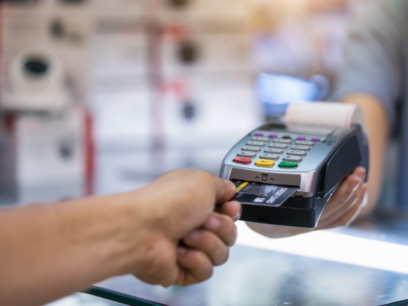Person with hand on their credit card, feeding it into a portable point of sale system, with a blurry background and receipt starting to be printed.