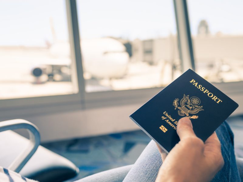 Person's hand holding a US passport with a plane in the background while sitting in an airport chair, unpainted nails, wearing jeans, looking out window at plane.