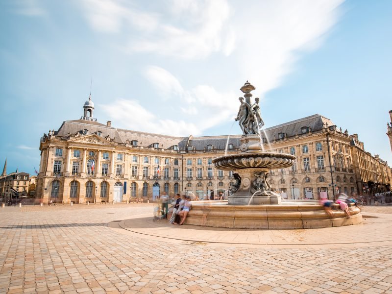 Fountain with people looking into it, clear street except for people at fountain, and a historic building in tan color behind the fountain in the heart of Bordeaux 