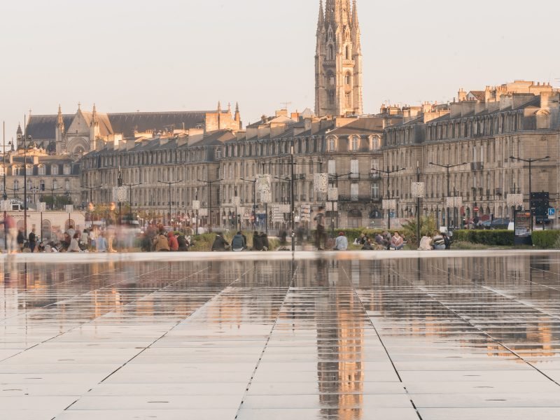 Rain slicked ground in the winter, with tower visible in the reflection on the wet ground, and other buildings and church in the distance, a few people sitting on steps in front of a green space, lamplights