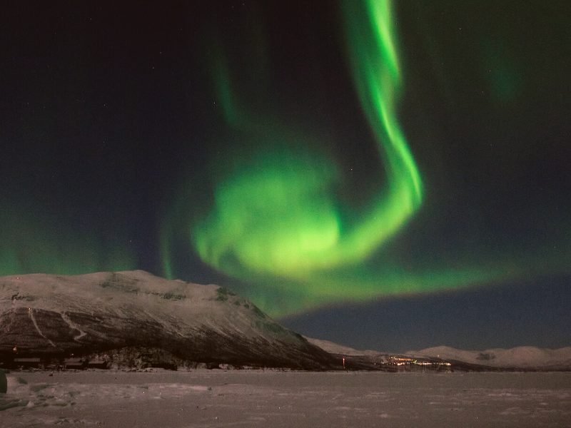 The neon green swirl of the Northern lights overhead with view of the landscape of Abisko and city lights in the distance.