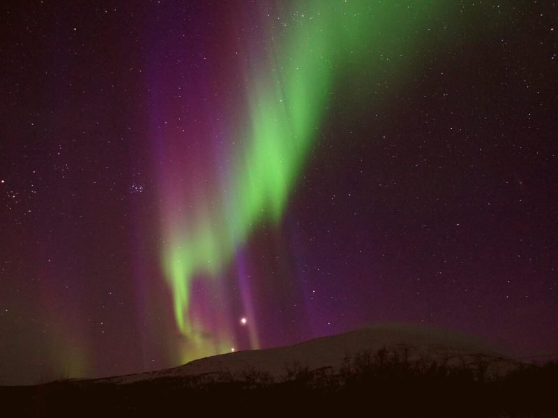 View of the aurora over the mountain near Abisko, Sweden, with green and purple colors in a zigzag shape against the sky.