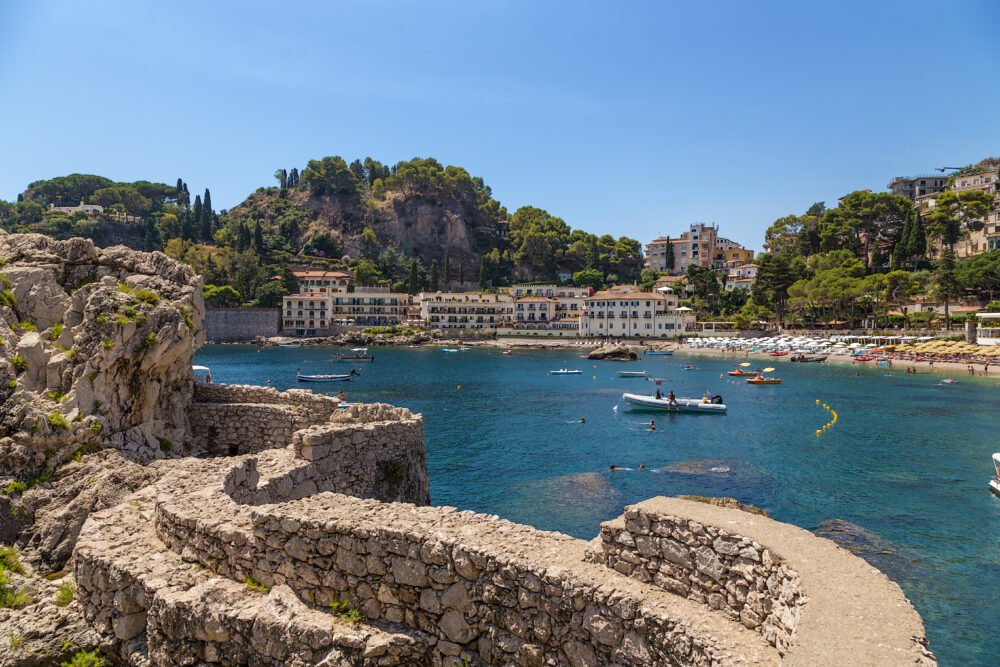 A stone wall looking onto the Gulf of Mazzaro in Taormina with beach chairs on the beach opposite the ledge