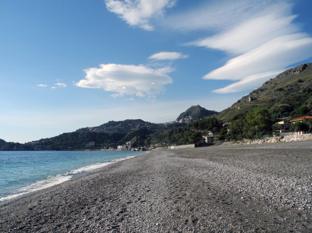 Black pebble beach in Sicily, a beautiful pebble beach near Taormina, with blue water and Sicilian hillside landscape.