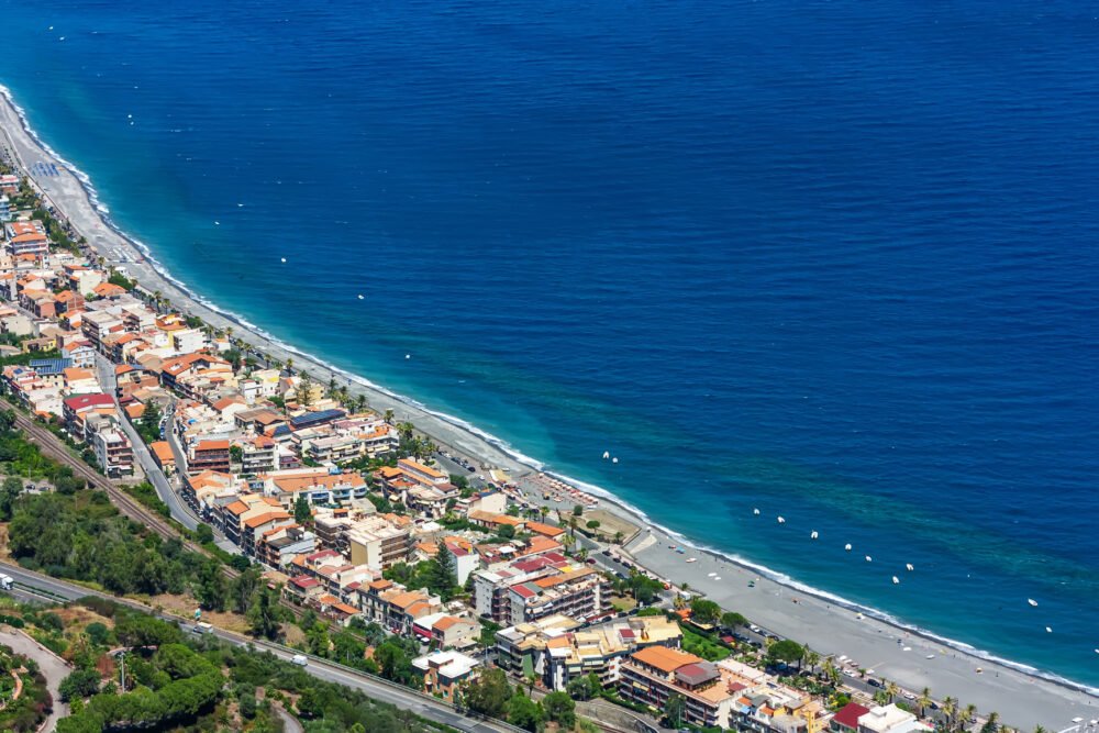 Gray-ish sand beach with blue, turquoise sea and houses around it, also roads.