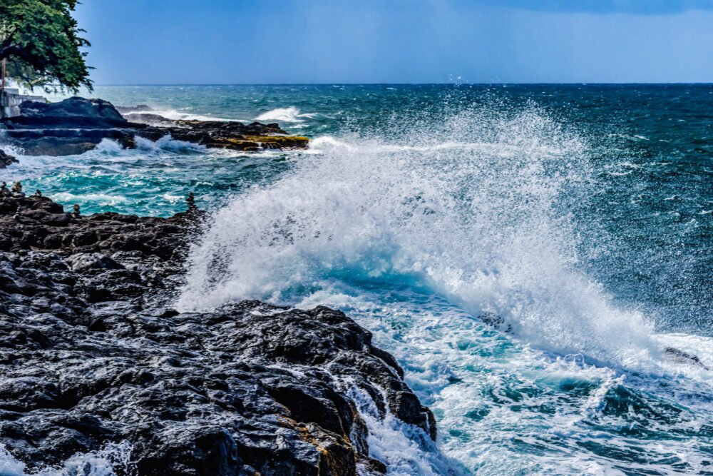 Black Lava Rocks with big waves, Blue Water erupting from a Blowhole which is like a marine geyser, ocean surf comes into stone lava tube and pushes water up through the hole