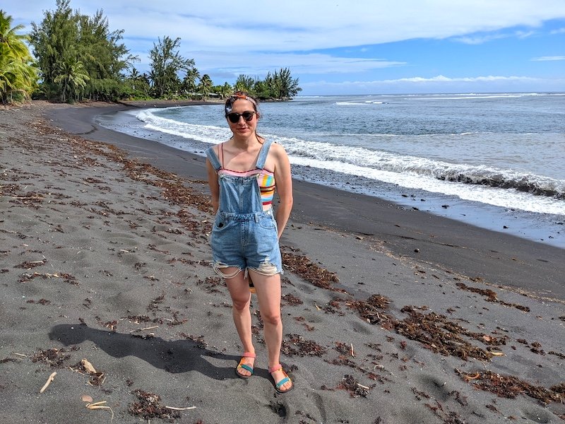 Allison standing on a black sand beach in Tahiti with overalls and a rainbow shirt on