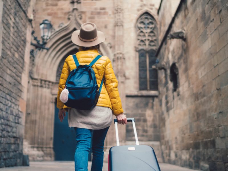 Woman with a brown hat, yellow jacket, sweater, jeans, backpack and rolling bag walking through the old quarter of Barcelona in December weather