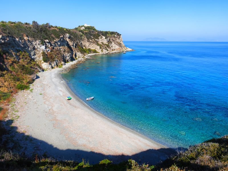 An isolated beach on a cove, with one boat visible, and otherwise the beach is entirely clear, with vibrant blue but still very clear water.