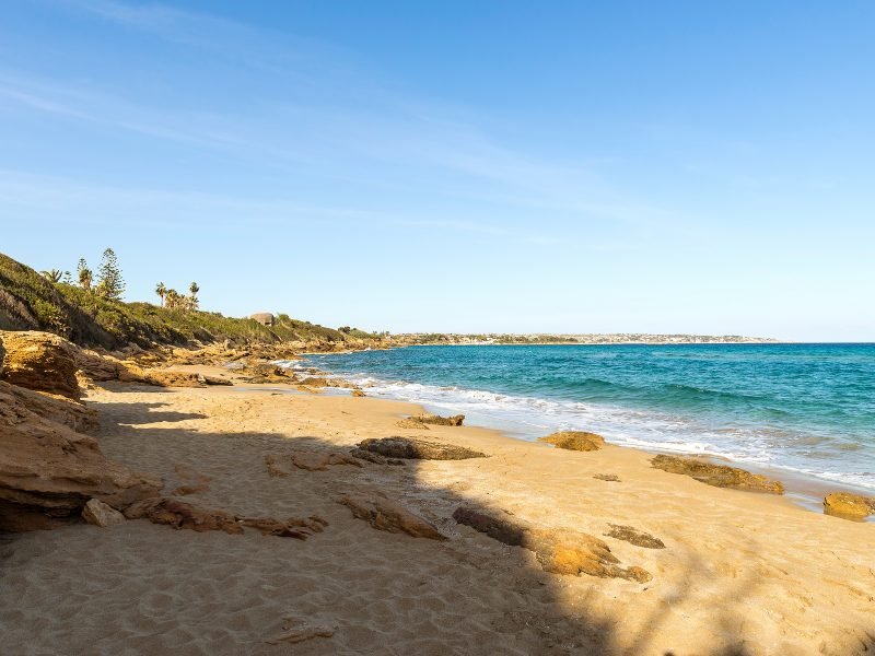 Sandy beach near Siracusa Sicily with rocks and sandy shore leading into turquoise water on a sunny, cloudless sky day in summer.