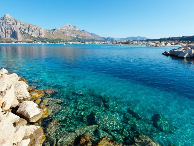 The shimmering, clear teal-blue water in Sicily's San Vito Lo Capo, with rocks in the foreground, and a mountain view in the background, and a town visible in the distance, all on a sunny summer day.