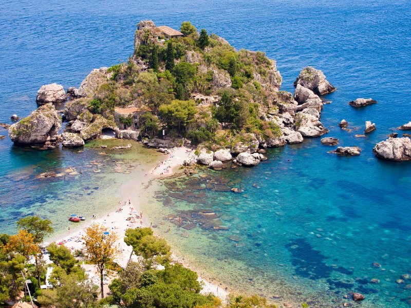 A small islet called Isola Bella (beautiful island) with a house atop the hill, a narrow strip of sand connecting it to the mainland, and people enjoying the sand from a view above on a hill overlooking the island and the cerulean blue sea.