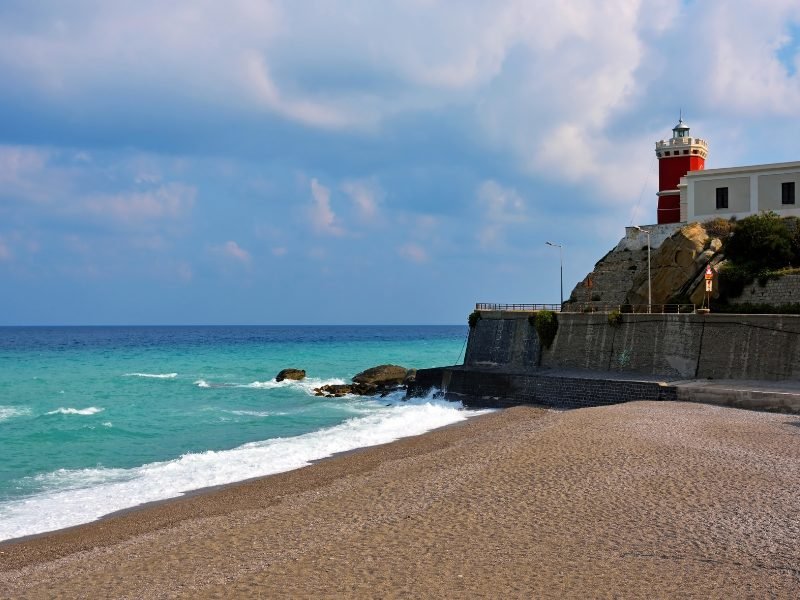 Sandy beach at Capo d'Orlando with turquoise sea crashing ashore, rocks, and a red lighthouse visible to the right of the frame.