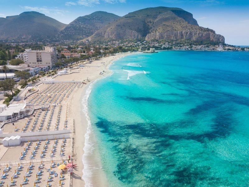The vibrant turquoise and cerulean colors of the water on the Sicilian coastline, with mountains in the background, and hundreds of beach chairs and umbrellas waiting for beach visitors to enjoy them.