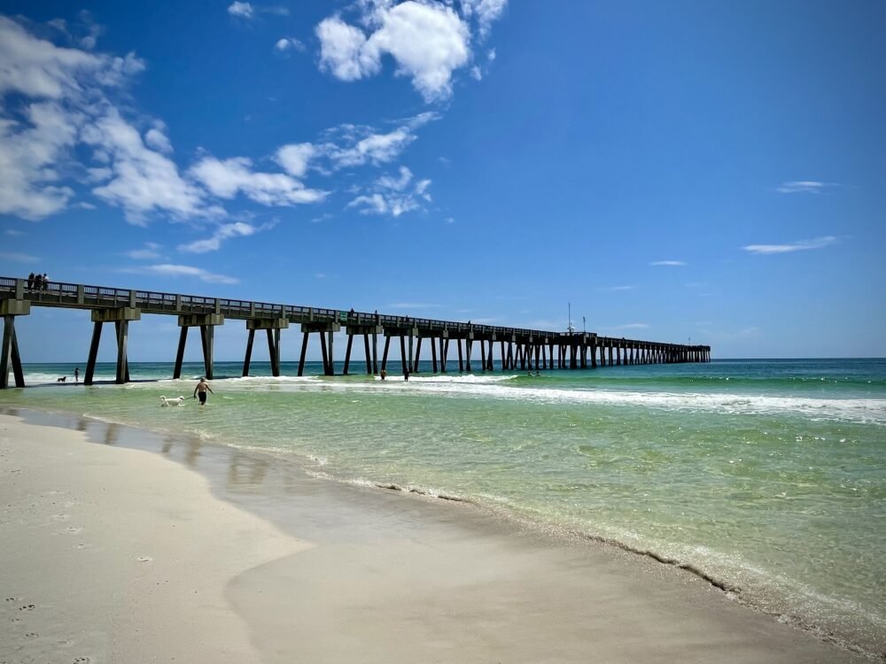 View of the beach and a pier leading out to the ocean on a sunny day with people enjoying the beach in Panama City Beach FL