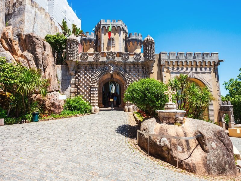 One way of entering the Pena Palace, with beveled architecture, flag of Portugal, archway and some people walking into the building