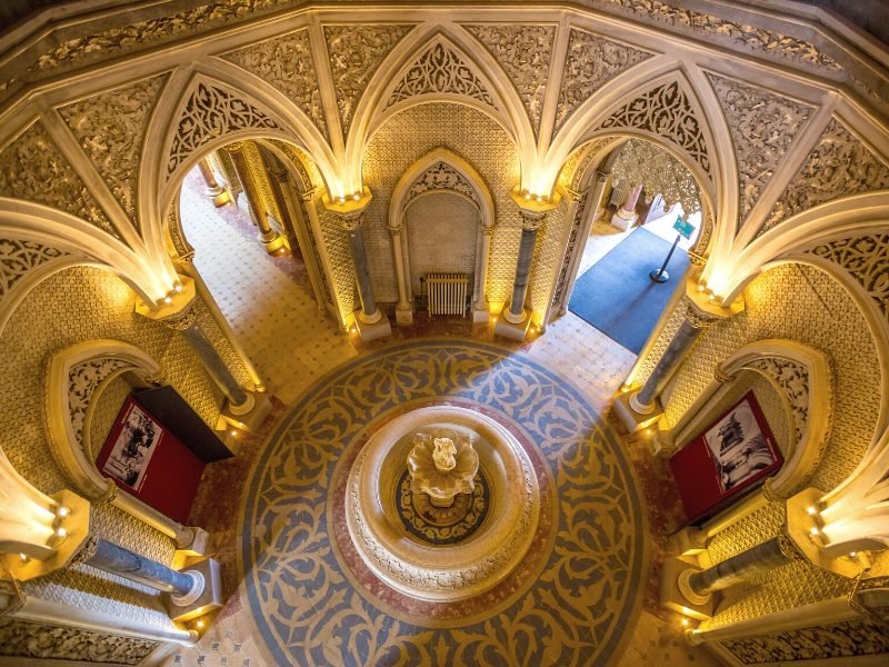 Ornate gold-toned interior of the Monserrate Palace in Sintra, with lots of archways and light streaming in through an open door, and mosaic floor