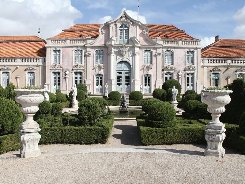 View of pink facade of Queluz palace outside of Sintra, portugal with symmetrical architecture and manicured garden with statues