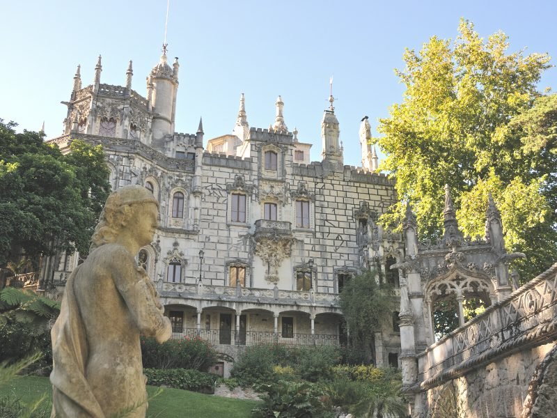 The gray stonework facade of the Quinta da Regaleira palace with lots of spires, architectural details, a statue in front as well as trees.