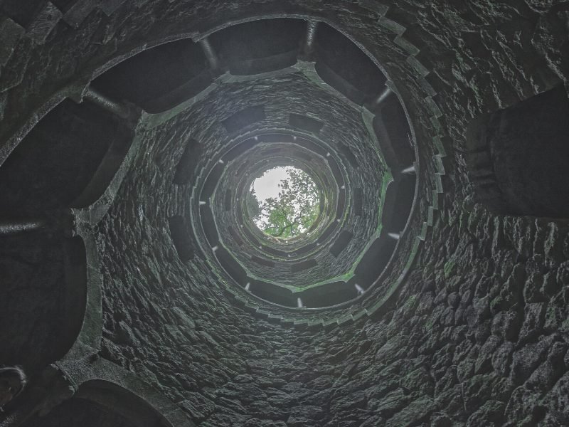 View from down below in the Initiation wells, looking up towards the sky, seeing some trees overhead as well as a spiral staircase built into the stone well