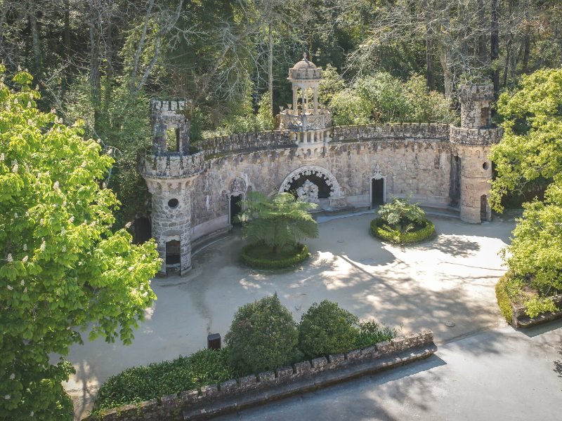 View of some of the grounds of Quinta da Regaleira, a popular landmark in Sintra, with a rounded arch stone walkway with three pillars amidst gardens