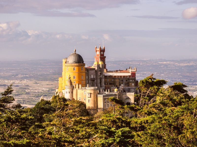 The red and yellow structure of the Pena Palace building in Sintra, near Lisbon, seen on a hill from a higher vantage point from afar.