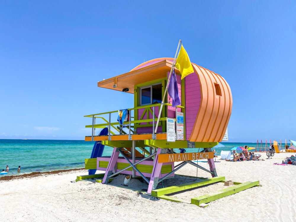 A colorful lifeguard tower that says Miami Beach located on white sand with turquoise and blue waters