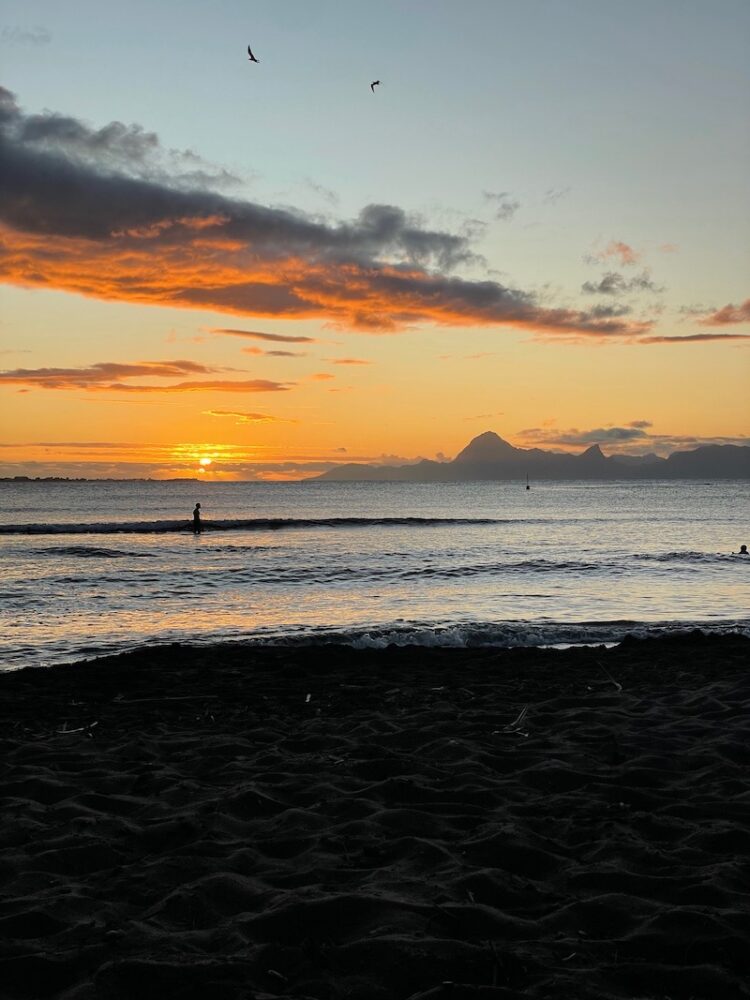 Black sand beach at Point venus with view of moorea at sunset with orange colors and sun setting on horizon