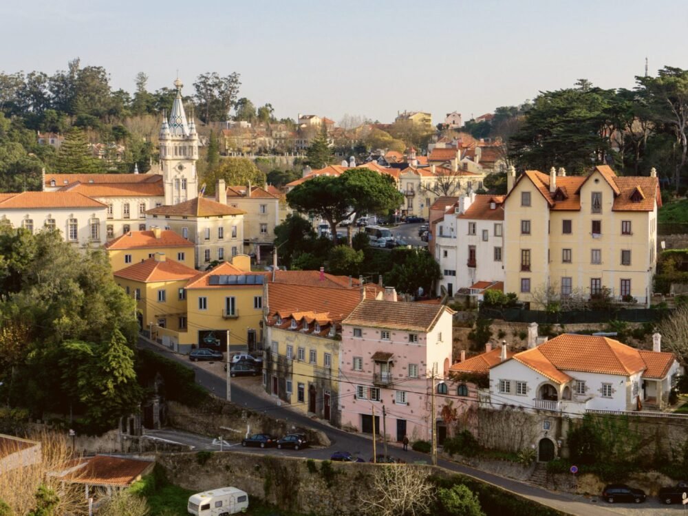 View of a road on a hill going down through the city center of Sintra, with old-fashioned buildings and a church or palace like building in the background.