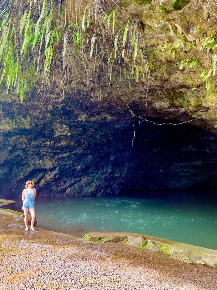 Allison standing in front of the grotto, which has ferns dripping down off the cave, with blue turquoise water underneath the cave, she is wearing a rainbow shirt and overalls and rainbow sandals.