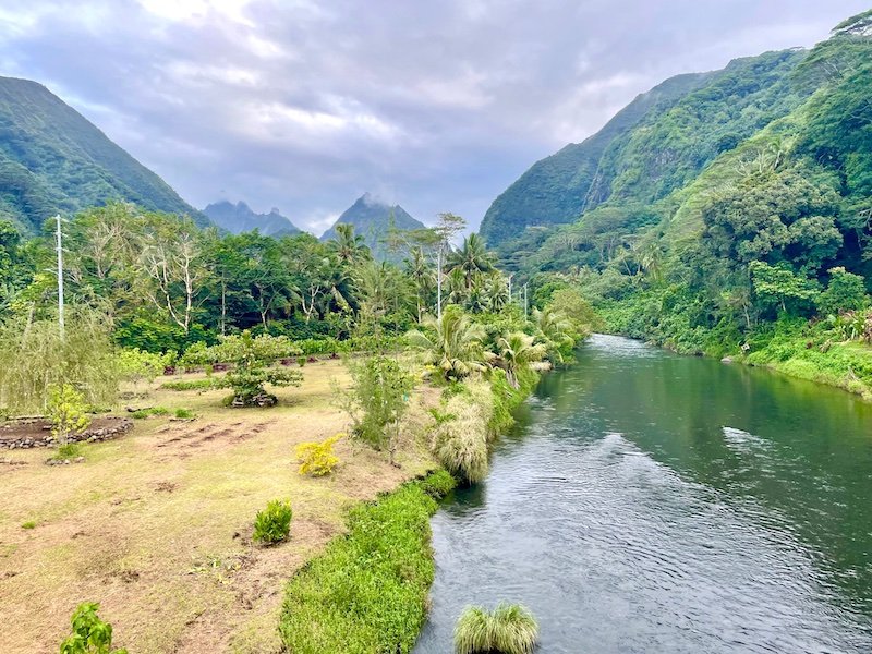 A river running through Tahiti Iti and leading out into the ocean, with palm trees and other lush green landscape and mountains in the distance, with a cloudy sky above.