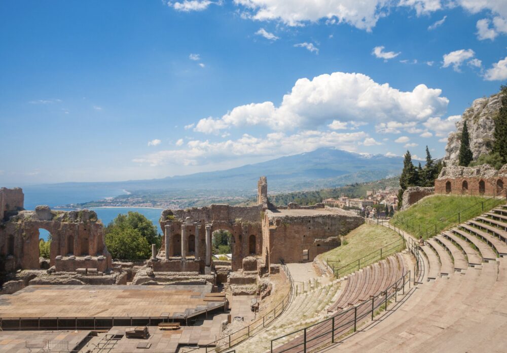 View of the beautiful Greek theater in Taormina with columns, stairs, amphitheater on a sunny day in Taormina, with Etna visible in the distance, as well as coastline.