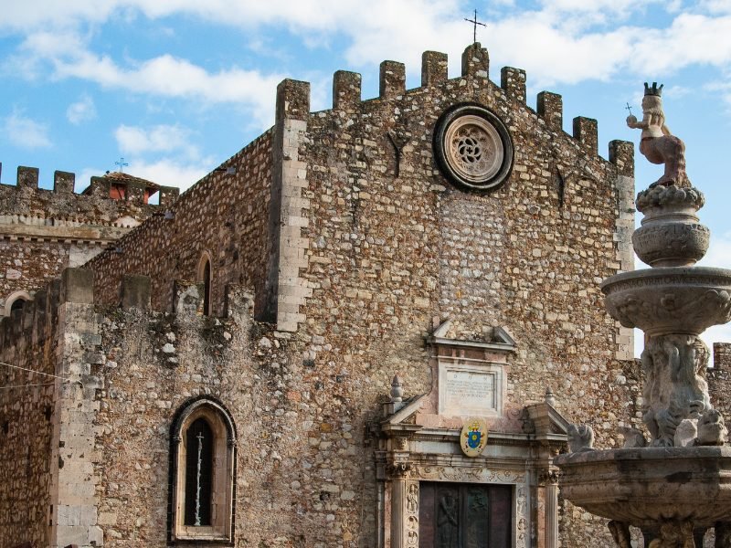 Stonework of the cathedral of Taormina, with an iron cross and a fountain in front of it