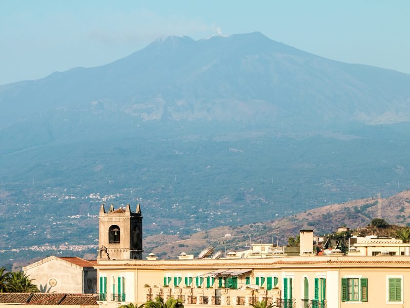 View of Mount Etna towering over the town of Taormina, with wisps of smoke or cloud visible on the top of the mountain