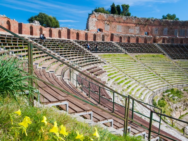 Yellow flowers blooming in spring at the Greek Theater in Taormina, with benches and rows of seats where some grass has grown in, and red brickwork from centuries past.