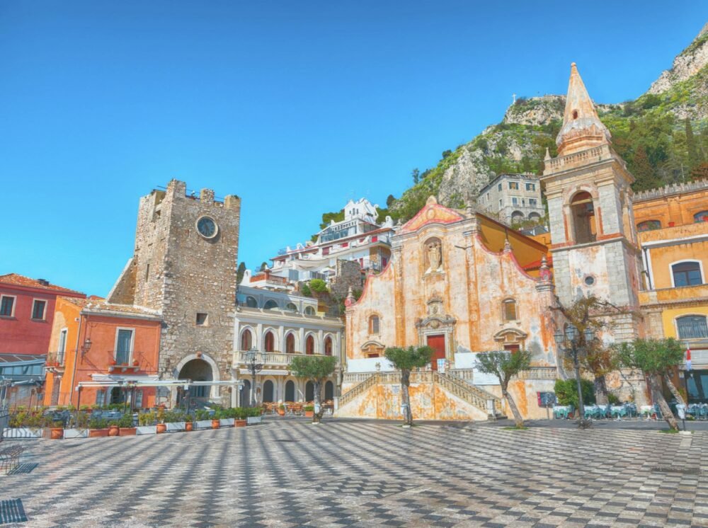 The central area of Taormina, with a yellowish fading facade church, a bell tower, and a clocktower in a stone tower, hillside behind it, and an empty piazza.