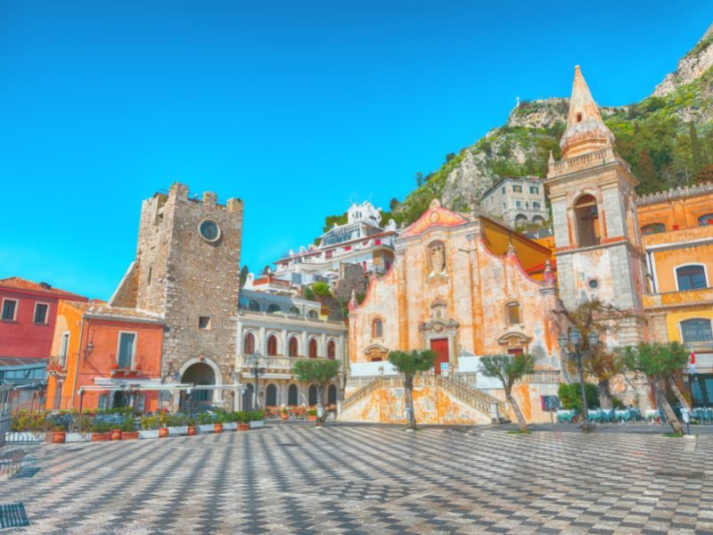 The beautiful checkered piazza ground in the city center of Taormina, with old tower, church, and hills in the background.