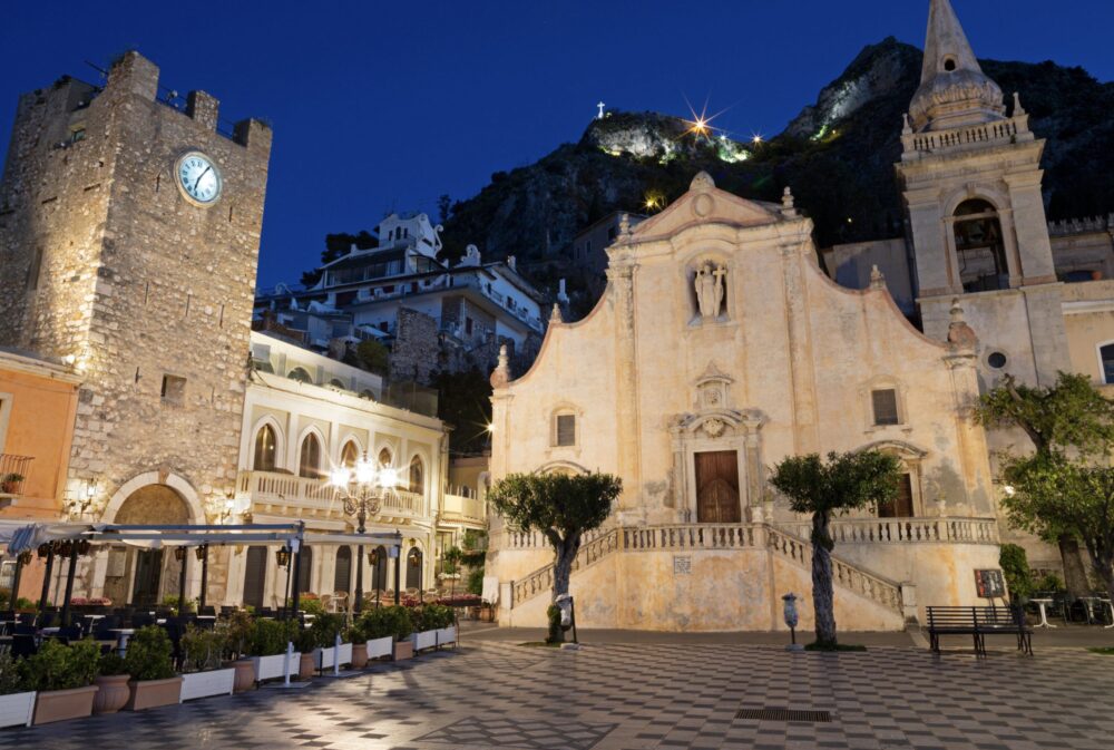 Nighttime view of Taormina's central piazza, bell tower, church, and castle on the hill above, with a small white cross, all seen at night.