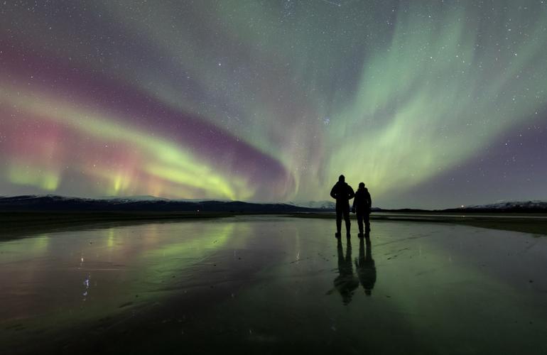 Two people standing on the frozen lake, looking at the aurora above which shows green, red, and purple colors swirling in the sky.
