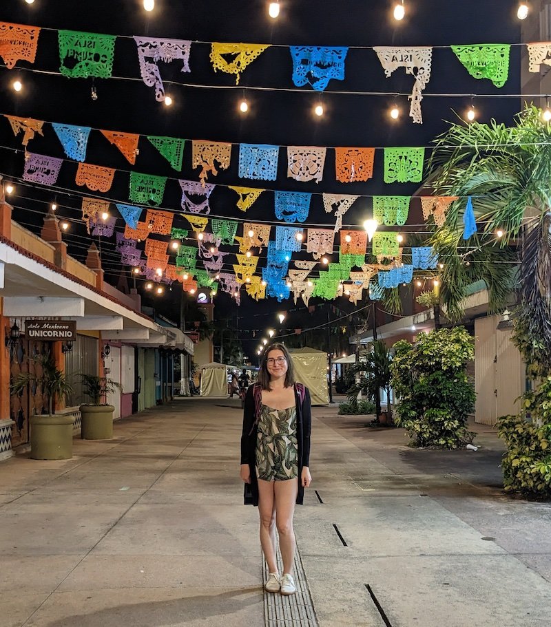 Allison Green wearing a plant green and white print romper and white tennis shoes and a long black cardigan on an empty street with colorful mexican-style paper flags strung above her, it is dark out and there is no one on the streets in Cozumel.