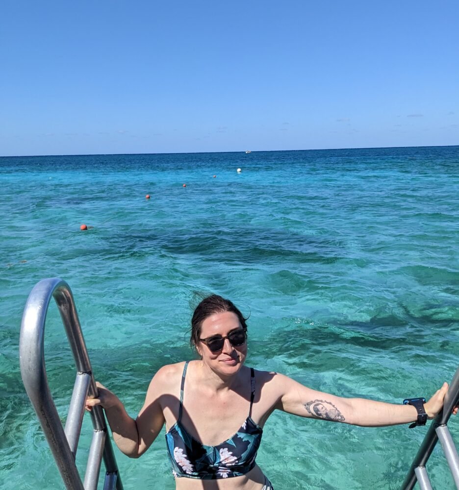 Allison Green, a white woman in a bathing suit with a tattoo on her inner arm, smiling at the camera while climbing up a ladder from the ocean in Cozumel