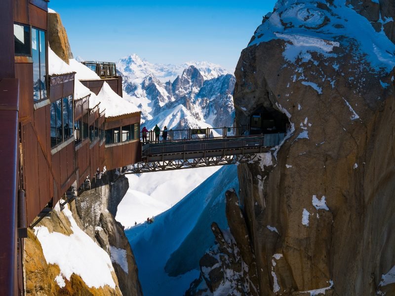 The mountain site of Aiguille du Midi in the winter with people standing on a bridge between a mountain refuge and a rock with a tunnel