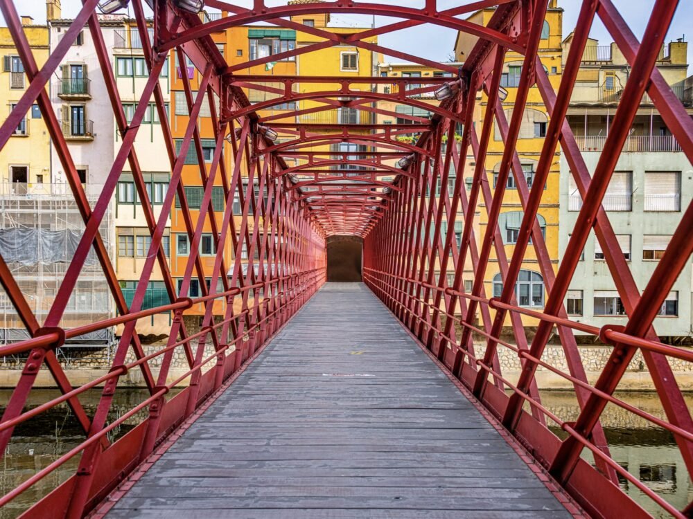 Red bridge with similar detailing to the Eiffel Tower, leading across the water to colorful buildings on the other side of the river