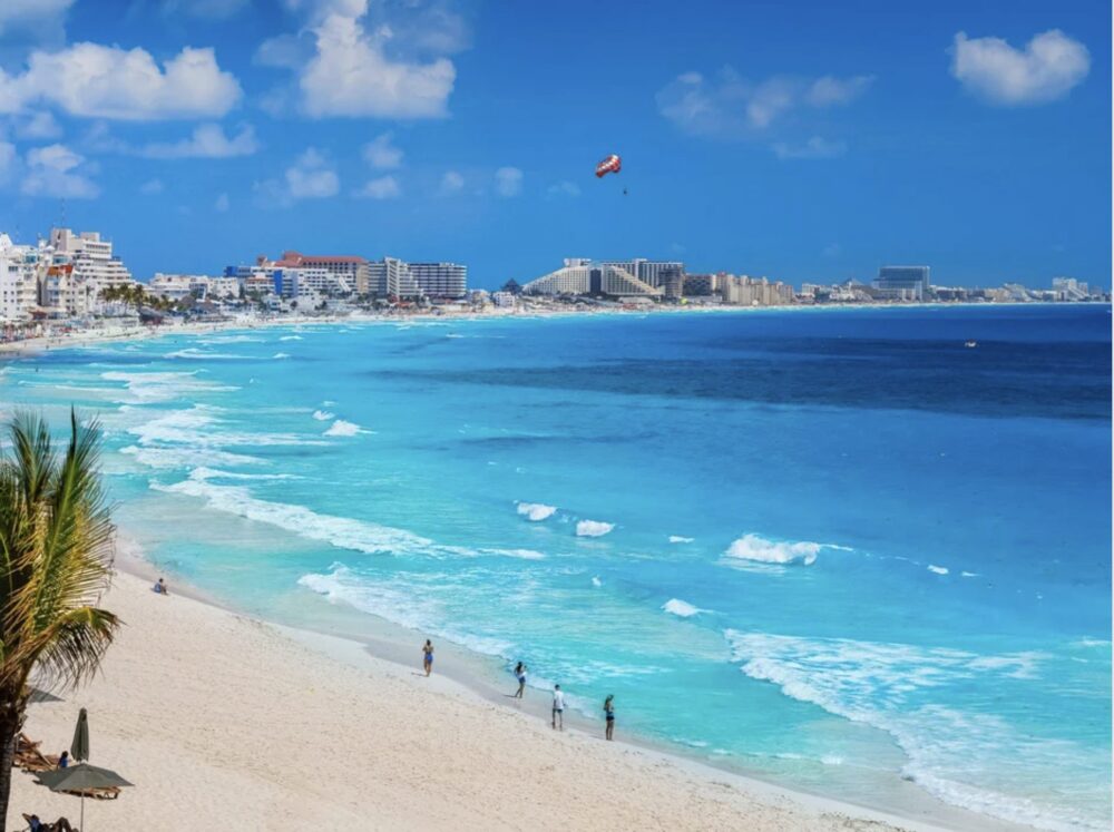 Beautiful bright blue water, leading out to dark blue water, in Cancun with a person parasailing over the Cancun skyline