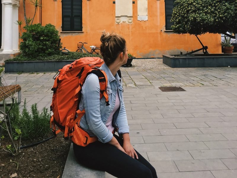 Woman sitting down in Italy wearing a large orange backpack with backdrop of orange-toned houses