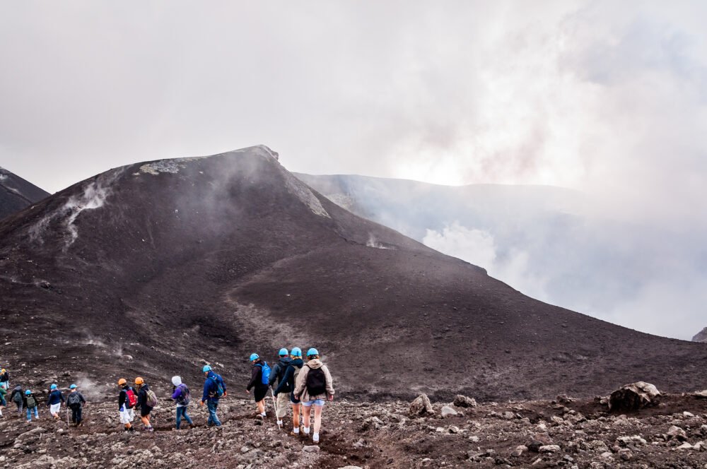 A group of travelers hiking to the summit of Mt Etna with helmets on and walking sticks