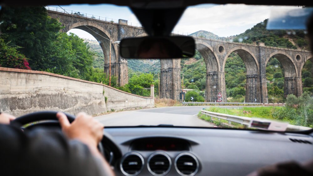 A person's hands holding the steering wheel while driving in Sicily near a giant aqueduct type structure